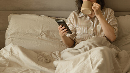 Woman drinking coffee and using phone in bed in the morning.
