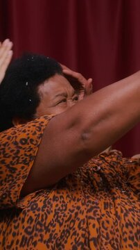 Vertical of delighted senior Black woman in leopard blouse with grandkids brushing colorful confetti from hair, laughing together inside cozy red-curtained photo booth after joyful celebration