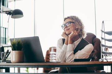 Young professional woman in office attire sits at her workplace, reflecting and smiling during a...