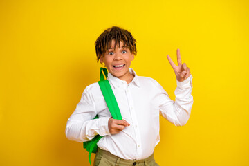 Cheerful boy in white shirt with green backpack showing a peace sign in front of a bright yellow...