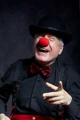 Portrait of a clown on dark background. An elderly man with red clown nose and bowler hat on head. Dressed in red pants and a black shirt with suspenders. The clown points to the side.