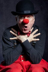 Portrait of a surprised clown against a dark background with hands on chest.