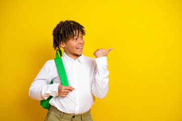 Smiling schoolboy with a backpack on a vibrant yellow background pointing sideways in a fun and...