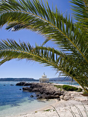 White-columned Greek lighthouse on Kefalonia’s rocky coast, framed by palm leaves and turquoise sea.