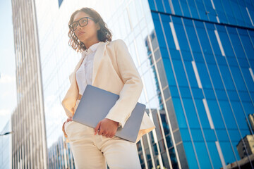 Elegant businesswoman holding a laptop, standing confidently outdoors, reflecting urban success and...