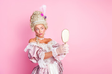 Elegant woman in historical costume admiring herself with a mirror, posed on a soft pink background.