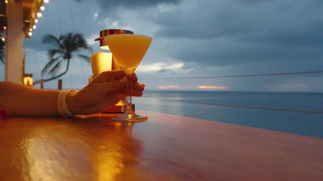 Woman Holding Yellow Cocktail in Martini Glass with Orchid Decoration at Oceanfront Bar During Sunset with Palm Tree and Ocean View, Tropical Vacation Concept