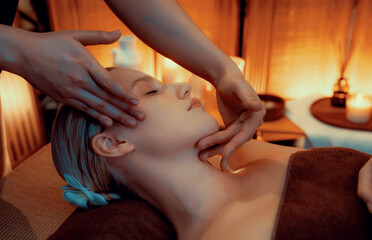 Caucasian woman enjoying relaxing anti-stress head massage and pampering facial beauty skin recreation leisure in warm candle lighting ambient salon spa in luxury resort or hotel. Quiescent