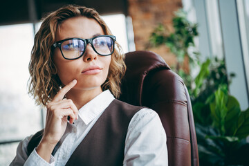 Confident businesswoman wearing glasses sitting in office chair, looking thoughtful and stylish,...