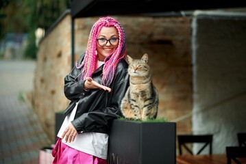 A woman with pink braids poses with a cat outdoors in a playful moment, both happy and relaxed.