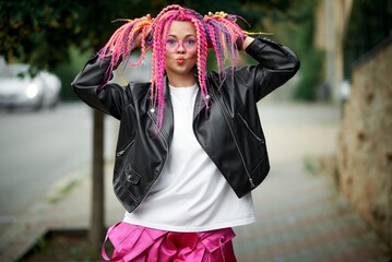 A woman with pink braids and glasses poses playfully in a black jacket outdoors.