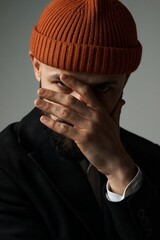 A stylish man wearing an orange beanie and black suit, hiding his face with his hand.