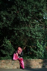 A stylish woman with pink dreadlocks poses outdoors near a brick wall with a tree backdrop.