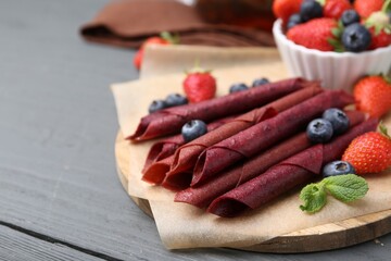 Tasty fruit leather rolls, strawberries, blueberries and mint on grey wooden table, closeup. Space for text