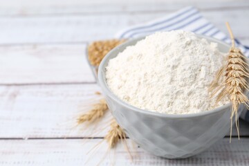 Wheat flour in bowl, grains and spikes on light wooden table, closeup. Space for text