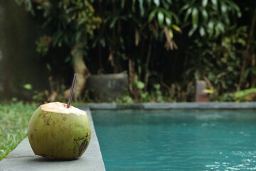 Green coconut with refreshing drink and straw near swimming pool outdoors, space for text