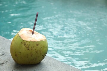 Green coconut with refreshing drink and straw near swimming pool outdoors, closeup. Space for text