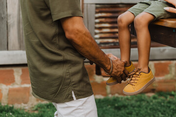Father tying his young son’s yellow sneakers while seated on a wooden bench
