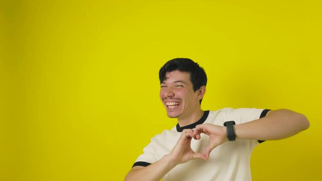 Very happy freckled young man in a white shirt making a heart with his hand with lots of energy on a yellow background with advertising space