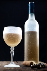 Crystal glass with white wine and bottle of wine served very cold,
over rustic wooden table on black background.