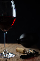 
Glass of red wine on the rustic wooden table, with corkscrew beside it and defocused wine bottle on black background.