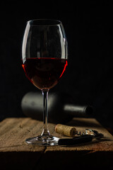 
Glass of red wine on the rustic wooden table, with corkscrew beside it and defocused wine bottle on black background.
