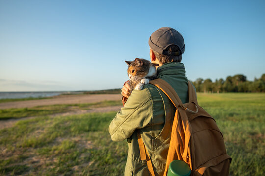 Man walking with cat in arms outside on beach. Curious pet looking around, exploring territory with interest, feeling cozy, comfortable in hands of male owner. Traveler wandering with kitty together 