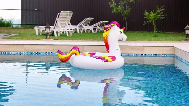 A playful inflatable unicorn float drifts in a resort pool with lounge chairs and green grass in the background, perfect for summer vacation.