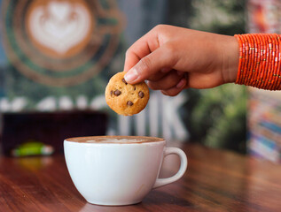 Woman dipping chocolate chip cookie in cappuccino at coffee shop
