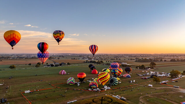 Hot air balloons float gracefully into the sky at sunset, creating a stunning visual display over a picturesque landscape filled with fields and spectators enjoying the vibrant event.