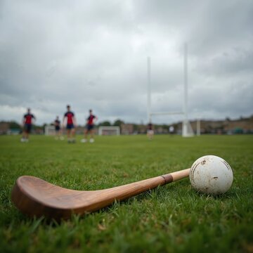 Wooden hurling stick, sliotar on green grass field. Players, goalpost in background. Cloudy sky. Traditional Irish sport, competitive game action, teamwork, healthy lifestyle. Gaelic athletic