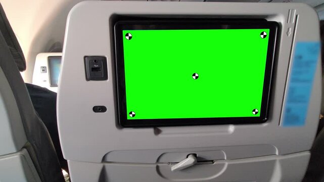 Close-up of an airplane seatback entertainment screen with green screen chroma key for video replacement, featuring a USB charging port, headphone jack, and seatback tray in a modern aircraft cabin