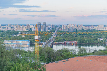 Overcast urban landscape of Kievs left bank, with a large body of water, boats, construction crane, apartment buildings, commercial structures, and soft even lighting
