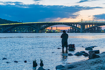 Person fishing near a metro bridge at dusk on the Dnepr River in Kiev Shallow water, rocky...