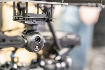 A combat quadcopter drone is shown, attached to a robotic arm in operation within an industrial workspace The arm moves from middle left to top right, with a spherical camera assembly at its end cap