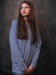 A woman with long hair is wearing a blue and white striped sailor shirt. She is standing in front of a dark background. Young model with slim body and rich long hair. Drees up party outfit
