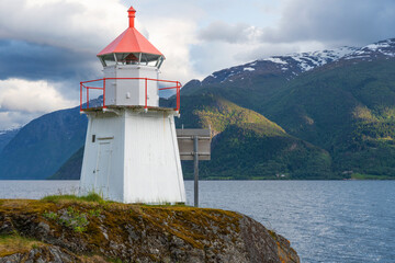 Small lighthouse on rocky hill along coast of vintage Norwegian town Leikanger along Norway's Sognefjord with blue skies small clouds and green snowcapped mountains in background