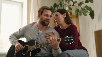 Happy couple in love dating. Man plays guitar to his girlfriend. Husband playing acoustic guitar for his wife at home. Anniversary. Enjoying music together. intimate moment. Playing guitar together - Powered by Adobe