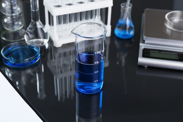 Beaker with blue liquid, other laboratory glassware and scale on black mirror table, closeup