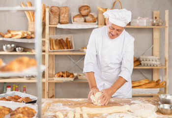 Elderly male baker in uniform kneading raw dough on table