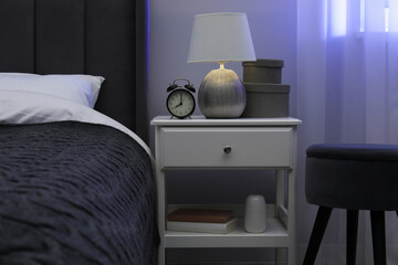 Bedside table with lamp, alarm clock and decor near bed indoors
