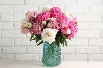 Bouquet of beautiful peonies in vase on white table against brick wall indoors