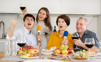 Happy family singing karaoke using microphone during family holiday, parents and adult children at festive table in light kitchen