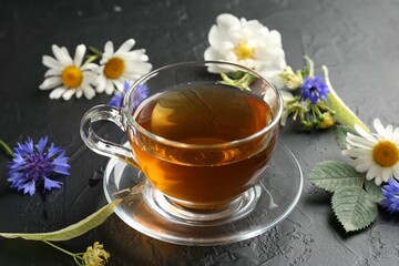 Aromatic herbal tea in glass cup, different flowers and green leaves on black table, closeup