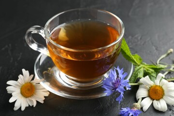 Aromatic herbal tea in glass cup, different flowers and mint on black table, closeup