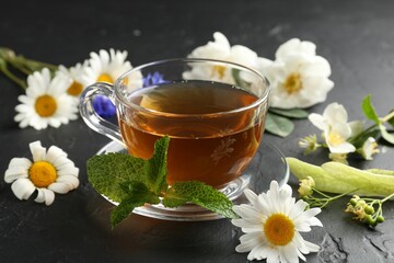 Aromatic herbal tea in glass cup, different flowers and mint on black table, closeup