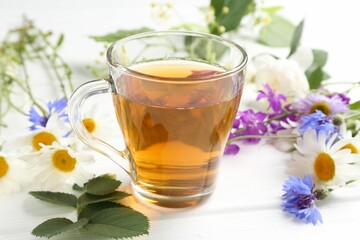 Aromatic herbal tea in glass cup, different flowers and green leaves on white wooden table, closeup