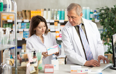 Obraz premium Elderly experienced pharmacist works at computer in a drugstore. Young assistant helps to find pills