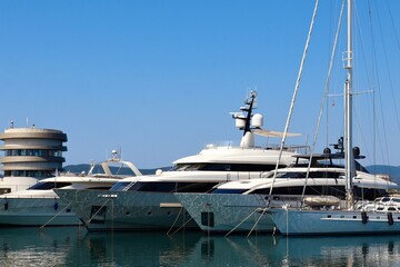 Fototapeta premium Luxury yachts are moored in a calm marina under a clear blue sky. The reflections of the boats shimmer on the water. A modern round harbor building is visible in the background.