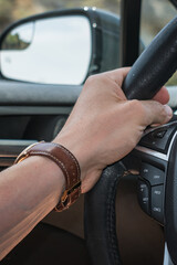 Male hand with watch and brown strap holding a car steering wheel